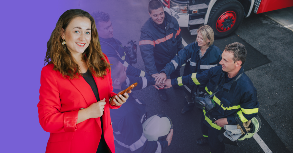 Osoba w czerwonej marynarce z telefonem, obok zespół strażaków dogadujących się w kręgu.